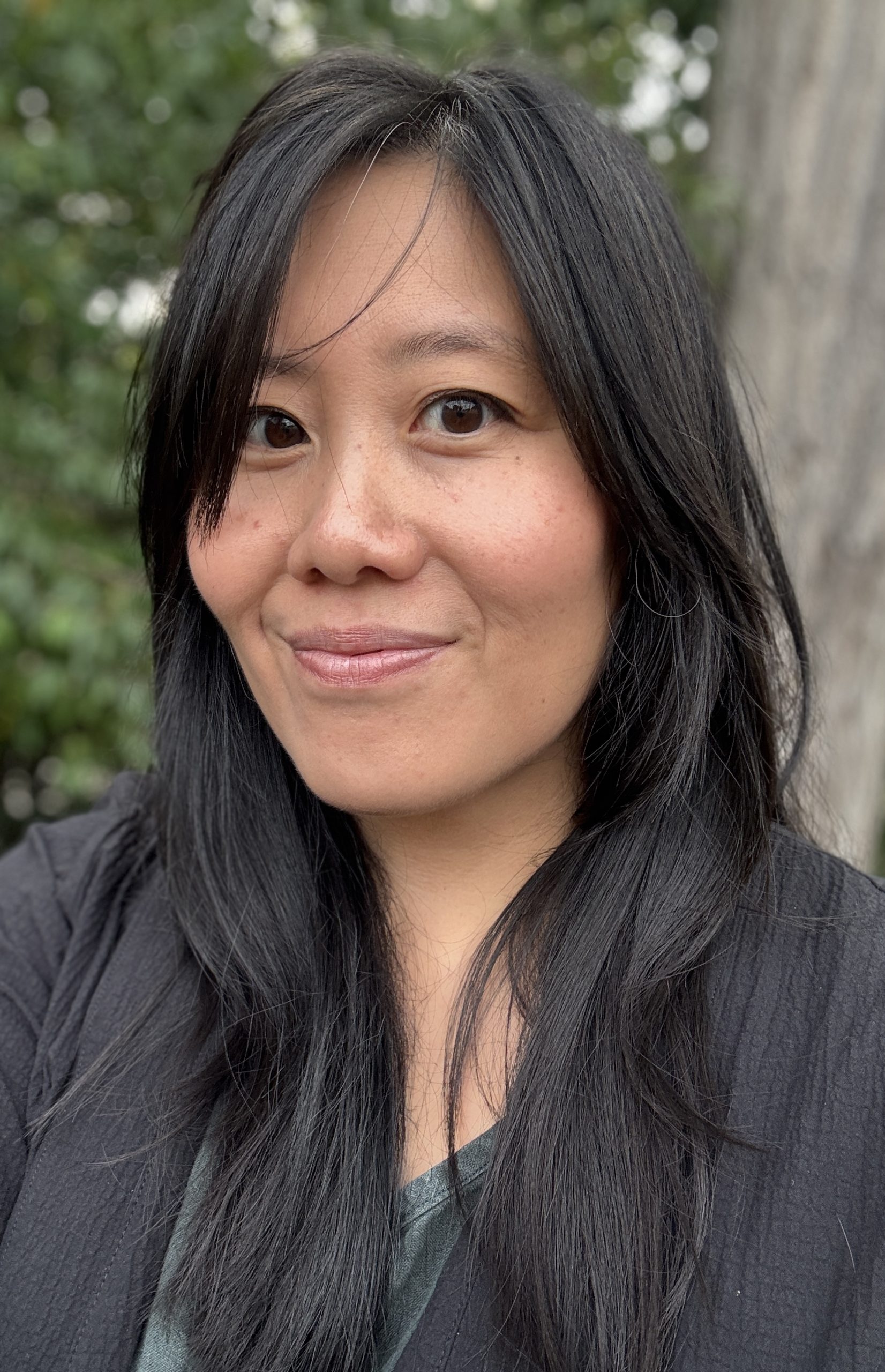 A person with long dark hair smiles at the camera, pictured from the shoulders up.