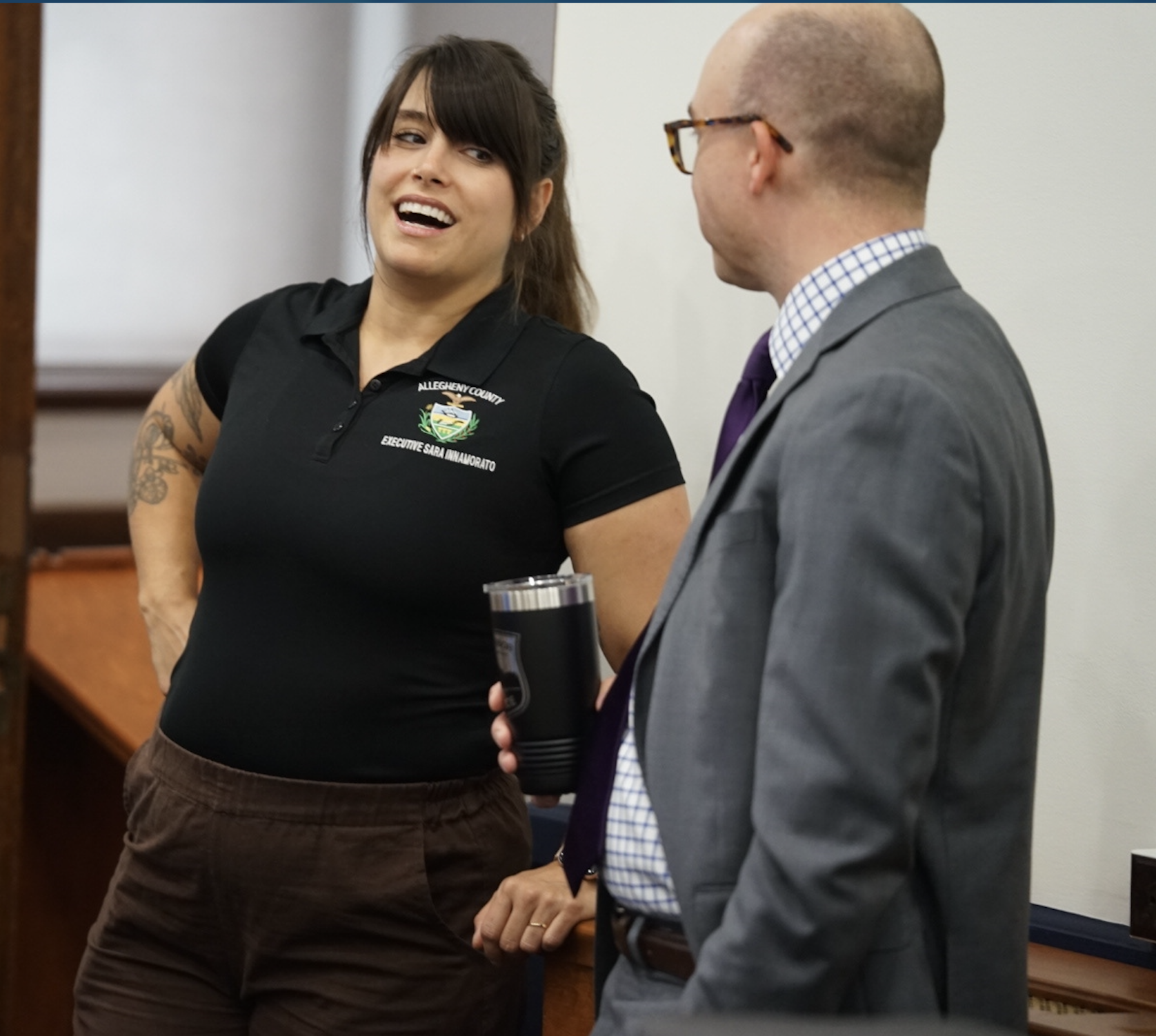 Allegheny County Executive, Sara Innamorato, in a black polo shirt talks to a man in a suit and glasses; she is smiling and holding a tumbler.