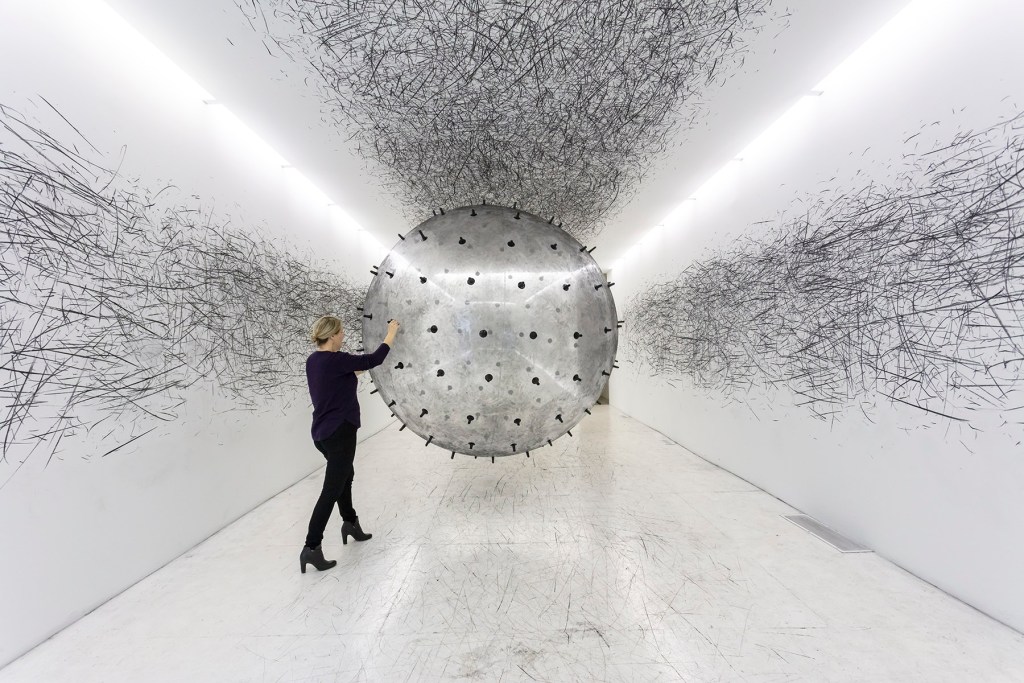 A woman in dark clothing pushes a clear ball with protrusions in a while room with charcoal marks on the wall.