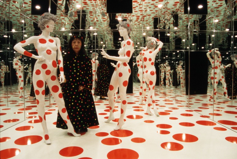 A person in a dark dress with polka dots stands in a room with mirrored walls and ceiling. There are mannequins in the room and they are covered with the same red polka dot pattern as the floor.
