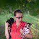 A woman in glasses holds a camera and a small dog wearing a dress while standing outside on grass with trees in the background.