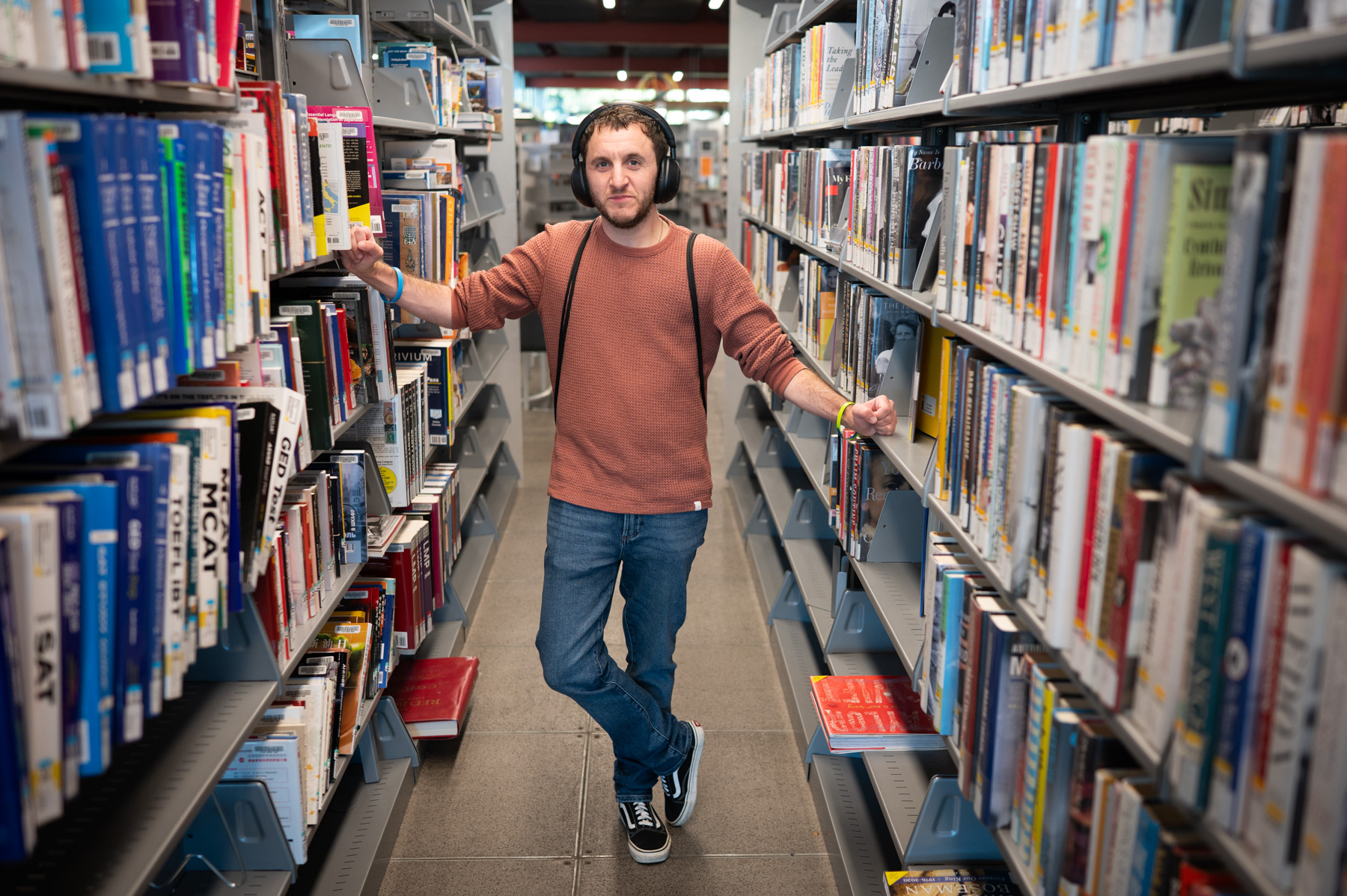 A man with headphones stands in a library, resting his arms on the bookshelves at either side.