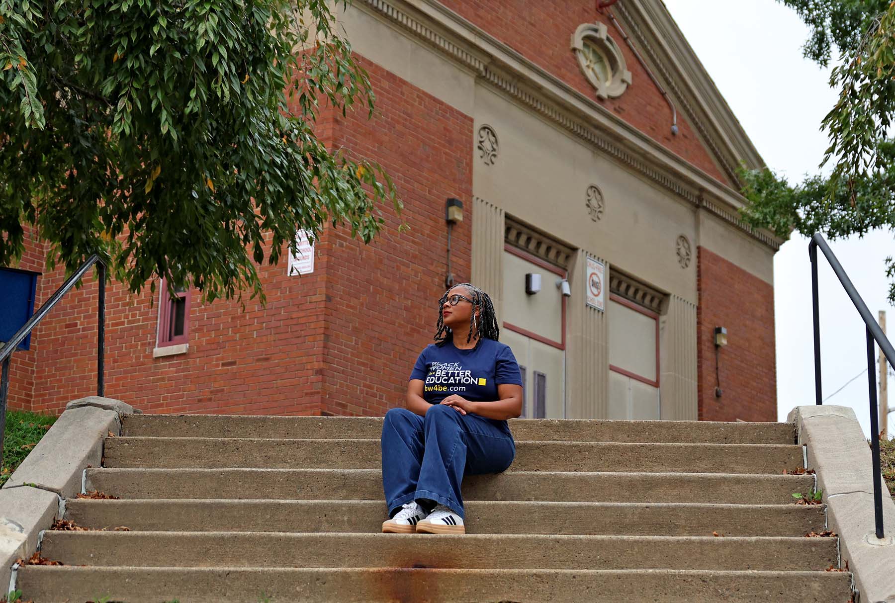 A woman wearing a dark blue t-shirt that says “Black Women for Better Education” sits on outdoor concrete steps in front of a brick building.