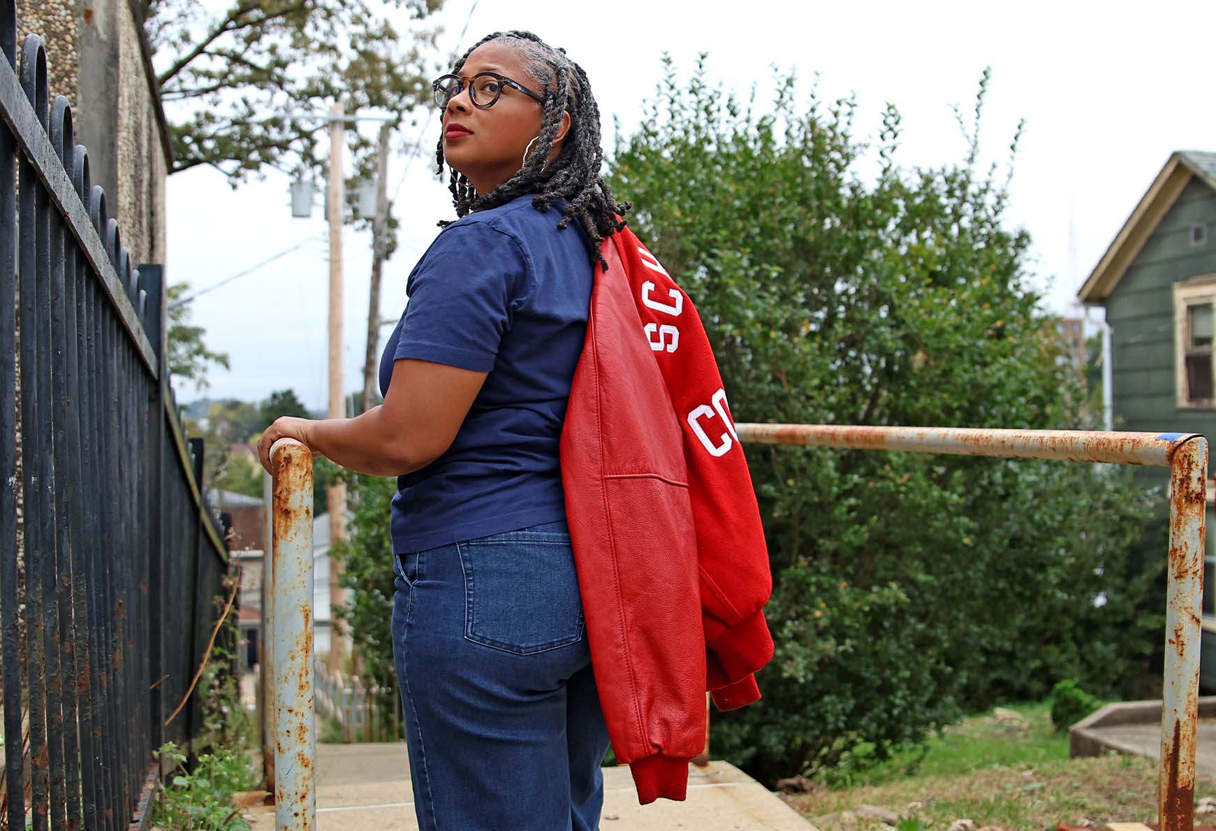 A woman with gray braids and glasses looks over her shoulder while holding a red varsity jacket over her arm.