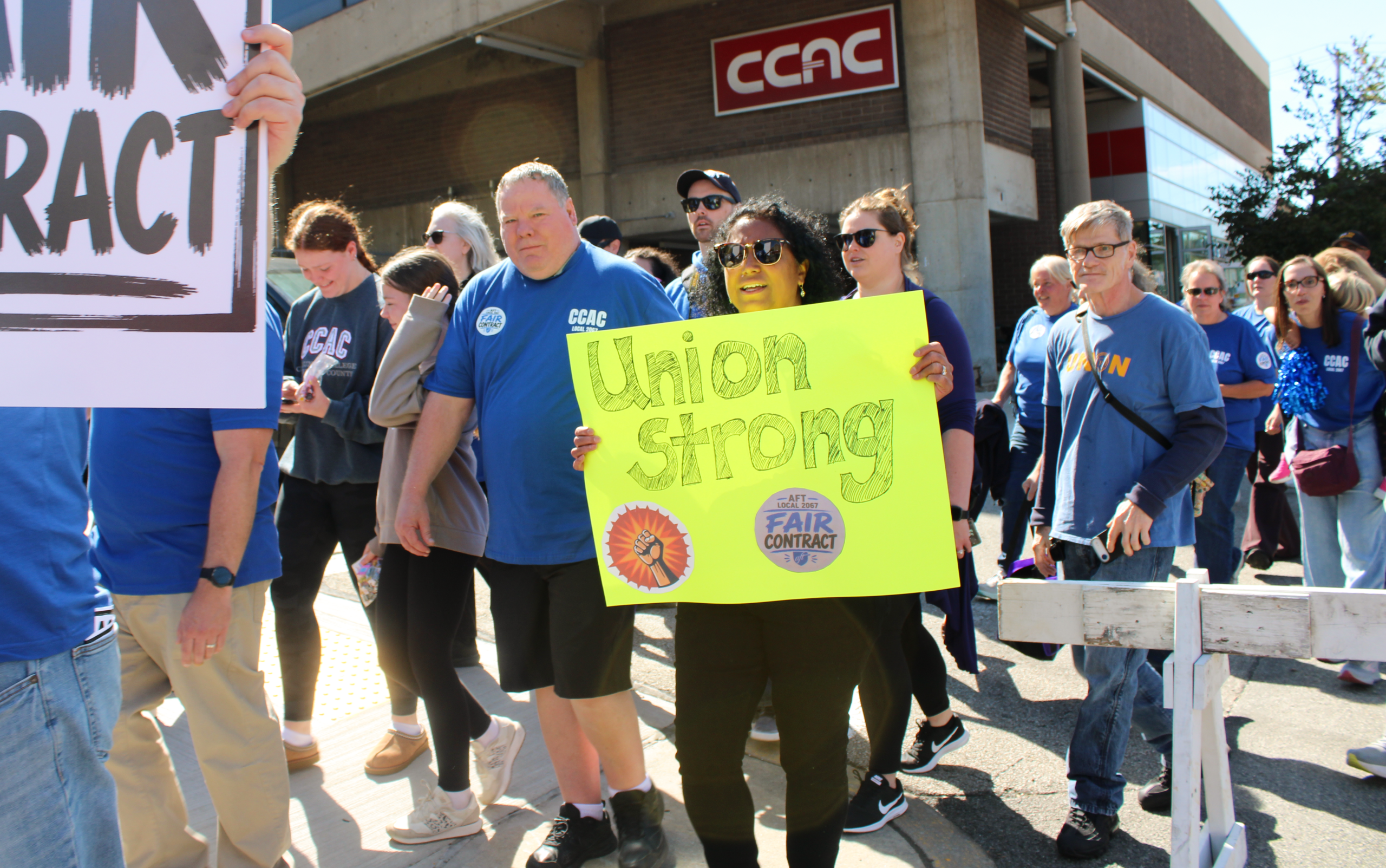 A group of people wearing blue CCAC shirts march while holding signs that read “Union Strong” and “Fair Contract.”